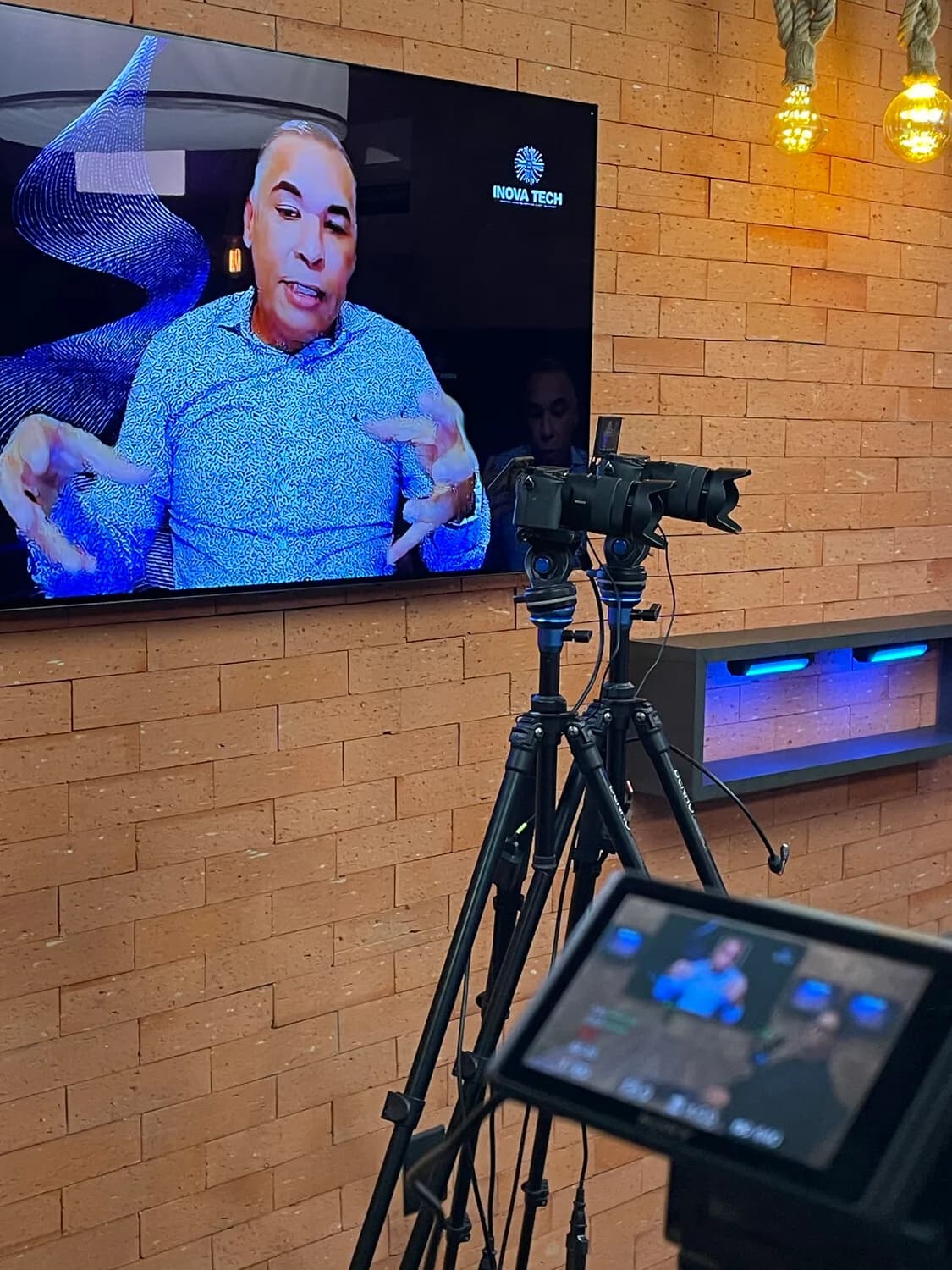 Câmera em foco com Joás Andrade desfocado ao fundo, monitor de transmissão